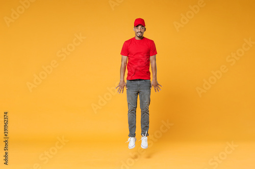 Full length fun delivery employee african man in red cap blank print t-shirt uniform workwear work courier dealer service on covid-19 virus concept jump run walk isolated on yellow background studio.