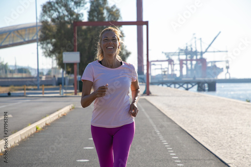 Happy excited mature woman jogging along river bank outside. Senior lady training for marathon. Front view. Activity and age concept © Mangostar