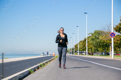 Focused mature woman in fitness clothes jogging along river bank outside  enjoying morning run. Front view  full length. Active lifestyle concept