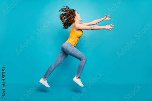 Fototapeta Naklejka Na Ścianę i Meble -  Funky wavy hairstyle lady jump high air spread arms wear casual yellow singlet jeans shoes isolated blue color background