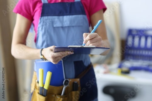 Woman builder in uniform writing with pen in document in workshop closeup. Provision of repair services concept