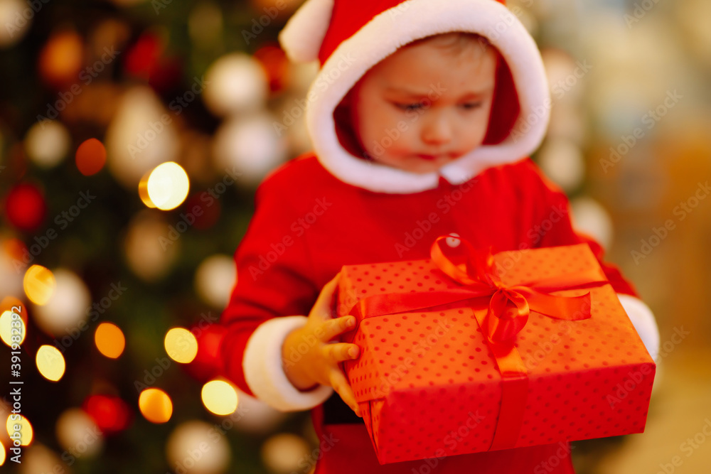 Christmas gift in the hand of Little child.  Kid in santa costume holding red present  box on the background of Christmas lights and tree. Winter holiday, New Year.