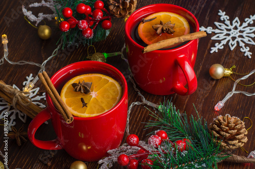 Aromatic, hot and delicious mulled wine with orange, cinnamon, star anise and cloves in red cups with Christmas decor. Feeling of celebration and warmth in the house
