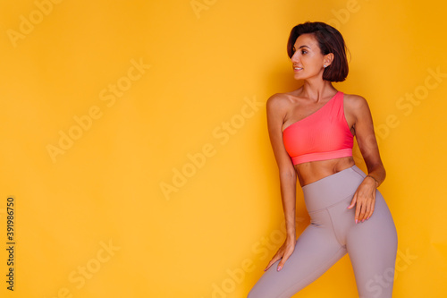 Sport and yoga lifestyle concept. Young fit pretty strong woman dressed in sport clothes, stylish top and leggings, poses against yellow background. Studio shot. 