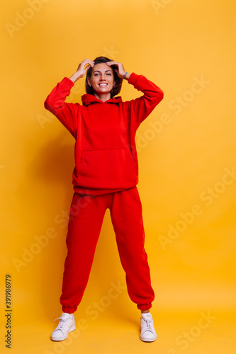 Portrait of happy tanned fit caucasian woman wearing trendy warm red fleece hoodie and pants, getting ready for cold winter. Studio bright shot, yellow background.  