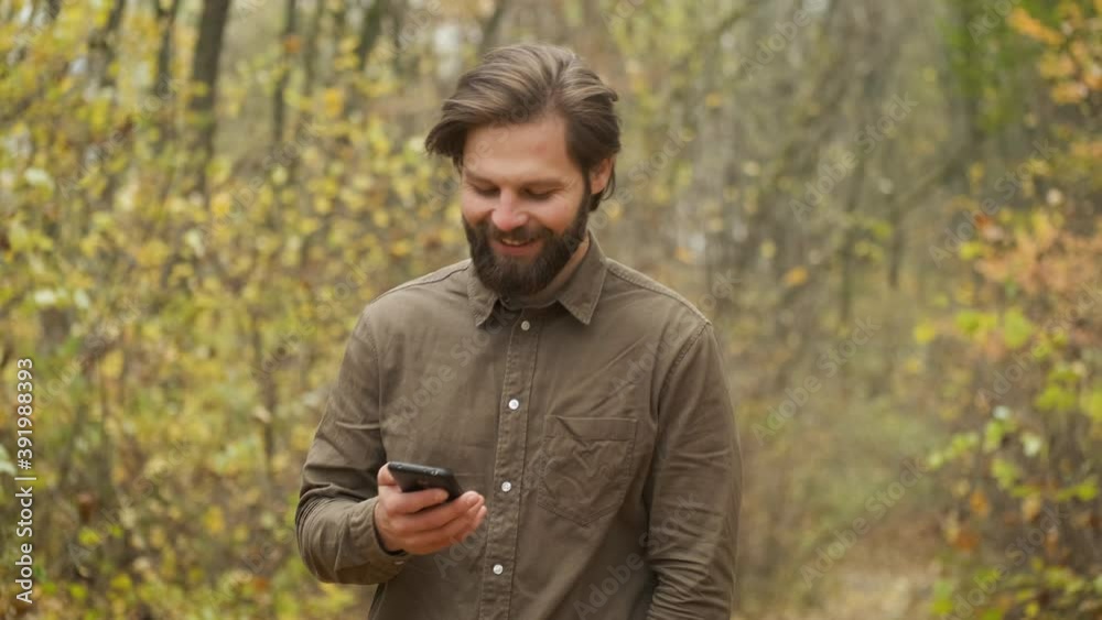 Smiling brutal man in a brown shirt walks in the autumn park holding a phone in his hands and communicates on social networks. Happy person