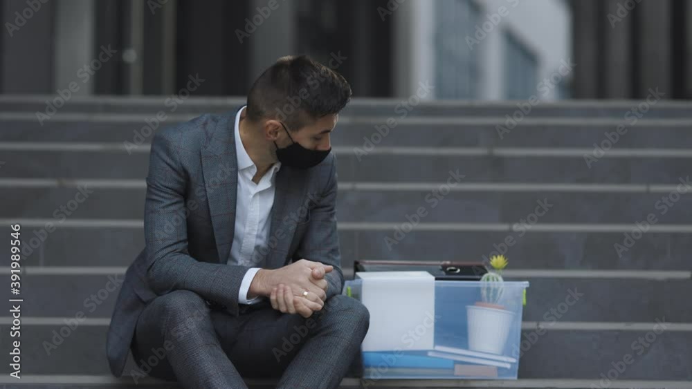 Unemployed businessman lost his business. Anxious concept. Workless man in despair. Fired male office worker in medical mask sitting on stairs in depression with box of stuff Unemployed businessman lost his business. Anxious concept. Workless man in despair. Fired male office worker in medical mask sitting on stairs in depression with box of stuff