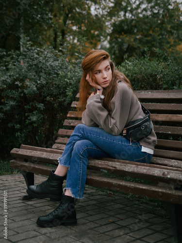Young thoughtful redhead woman in hoodie hand supports the chin enjoying warm fall day sitting in city park with green and yellow foliage background © sergeyzapotylok