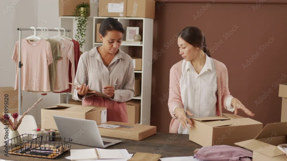 Medium shot of multi-ethnic young women working in office of small online clothes store and preparing packages for delivery. They are checking boxes and writing something on clipboard Medium shot of multi-ethnic young women working in office of small online clothes store and preparing packages for delivery. They are checking boxes and writing something on clipboard
