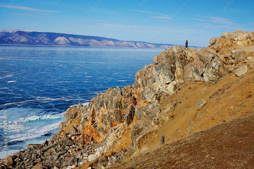 A man stands on top of the rocky Cape of Olkhon island and admires the vast expanses of frozen lake Baikal. Clear transparent ice with cracks. Travel in Russia in winter. A man stands on top of the rocky Cape of Olkhon island and admires the vast expanses of frozen lake Baikal. Clear transparent ice with cracks. Travel in Russia in winter.