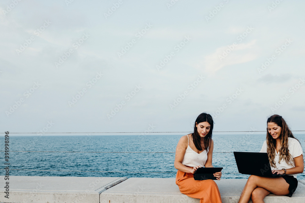 two women work with their computers and chat with the sea in the background