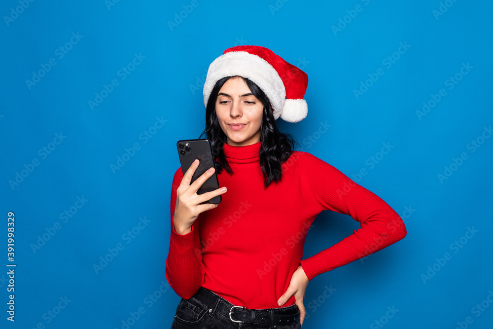 Beautiful woman typing on the phone with christmas hat is posing isolated on blue background. Beautiful woman typing on the phone with christmas hat is posing isolated on blue background.