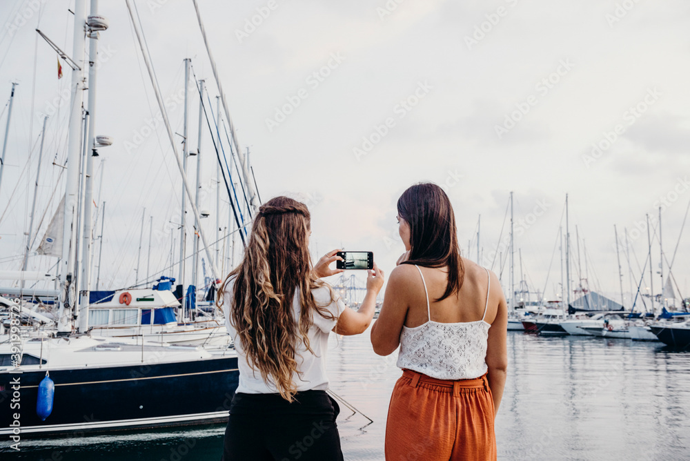 two women from behind take a photo with their mobile phone in the port