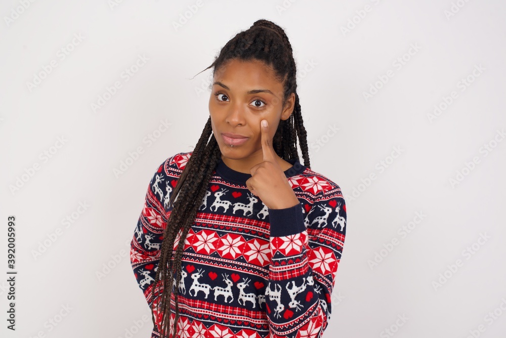 Young beautiful African American woman wearing Christmas sweater against white wall, looking, observing, keeping an eye on an object in front, or watching out for something. Young beautiful African American woman wearing Christmas sweater against white wall, looking, observing, keeping an eye on an object in front, or watching out for something.
