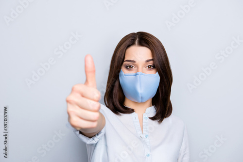 Portrait of brunette girl in blue fabric face mask gesturing thumb up symbol front to the camera isolated on grey background © deagreez