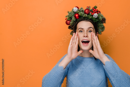Cute young woman in traditional Christmas wreath, openes mouth wide, screams joyfully, Xmas is coming, isolated on orange background with copy space. Happy New Year celebration merry holiday concept photo