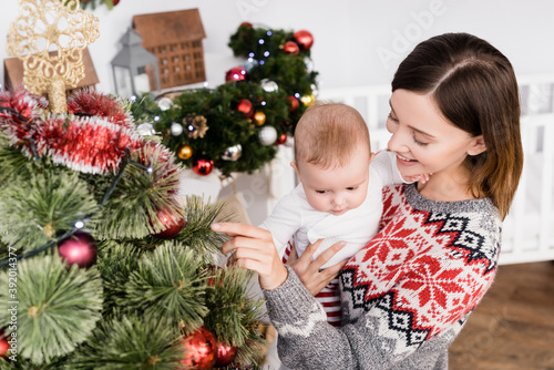 smiling woman holding in arms infant boy near decorated christmas tree
