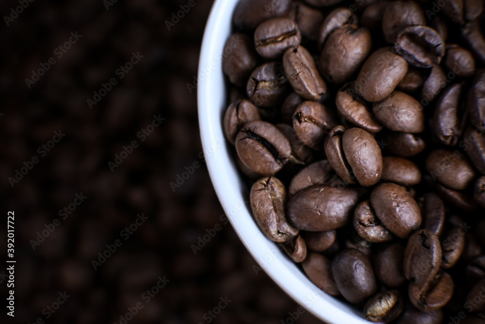 White cup full of coffee beans close up against the coffee beans background. Coffee mug. Morning espresso. White cup full of coffee beans close up against the coffee beans background. Coffee mug. Morning espresso.
