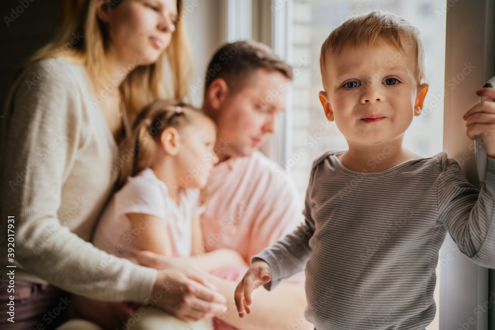 cute caucasian little boy standing on window sill. His parents and sister on background looking out the window. Home isolation during world pandemia. Image with selectie focus and toning cute caucasian little boy standing on window sill. His parents and sister on background looking out the window. Home isolation during world pandemia. Image with selectie focus and toning