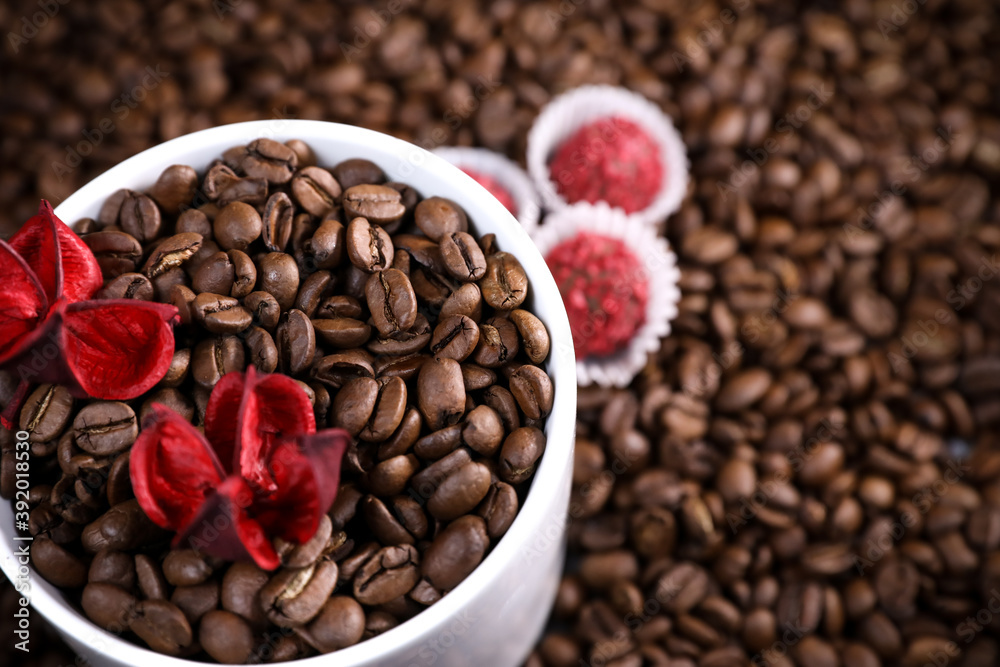 Fototapeta premium White cup full of coffee beans on a coffee beans background with red chocolate candy and red flowers. Morning espresso. Coffee mug.