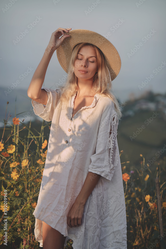 The blonde in a linen dress and straw hat walks among herbs and wildflowers on a meadow in the mountains. A woman in the Boho style, resting in the countryside, a simple village lifestyle