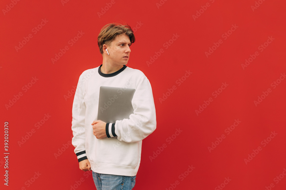 A handsome guy in casual clothes in wireless headphones and a laptop in his hands walks down the street against the background of a red wall and looks away at an empty place A handsome guy in casual clothes in wireless headphones and a laptop in his hands walks down the street against the background of a red wall and looks away at an empty place