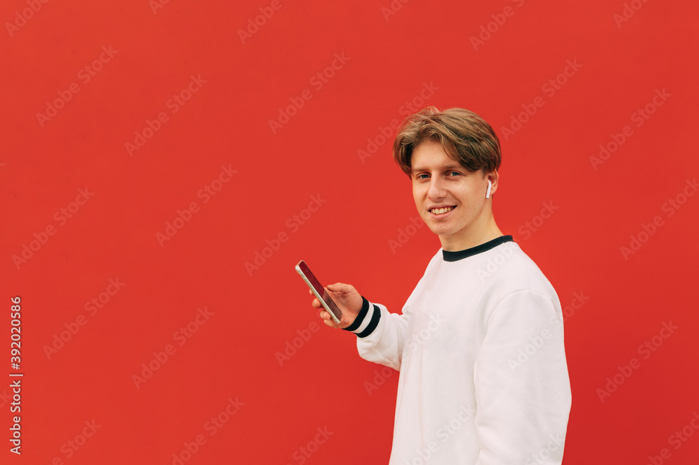 Portrait of happy guy in stylish casual clothes standing with smartphone in hands and wireless headphones in ears on red background and posing at camera Portrait of happy guy in stylish casual clothes standing with smartphone in hands and wireless headphones in ears on red background and posing at camera