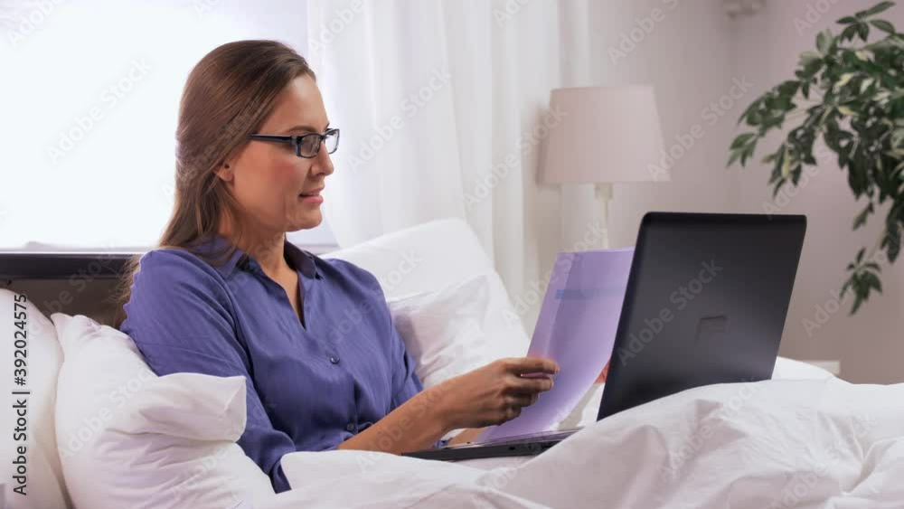 remote job, people and business concept - young woman in glasses with laptop computer and papers in bed working at home remote job, people and business concept - young woman in glasses with laptop computer and papers in bed working at home