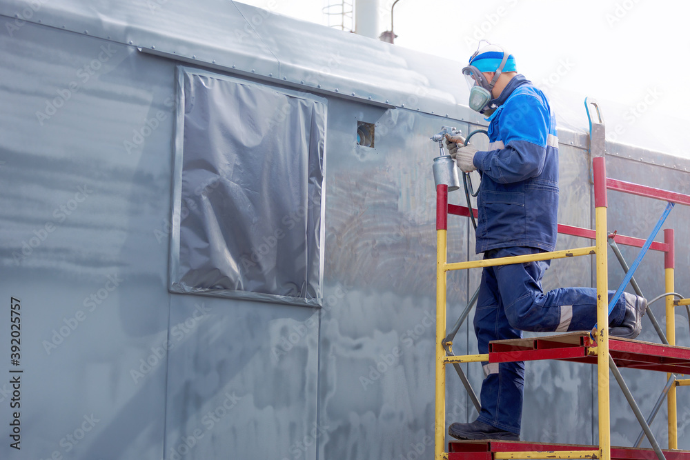Industrial work. Priming of metal products from the compressor gun. A worker in overalls and a respirator paints the body of a truck trailer or a metal car.