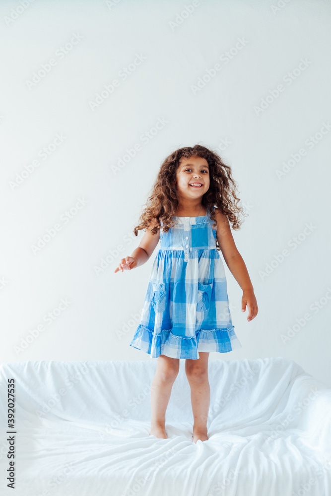 Portrait of a beautiful little curly girl in a blue dress on a white sofa Portrait of a beautiful little curly girl in a blue dress on a white sofa