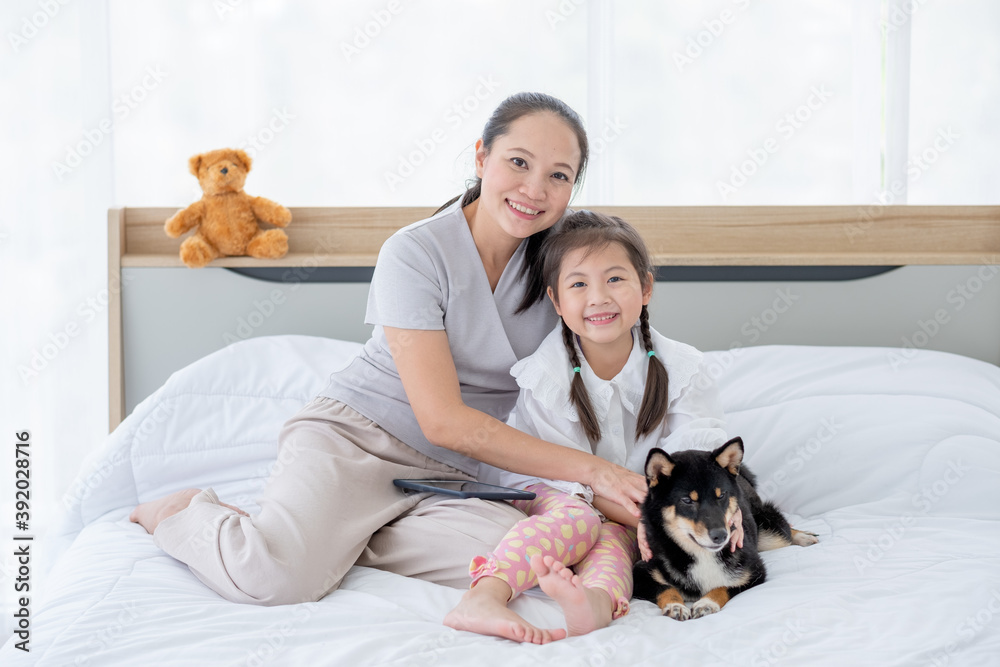 Asian mother and girl sit on white bed with black Shiba dog and look to camera with smiling in bedroom. Concept of happy family feel relax to stay home with their own pets