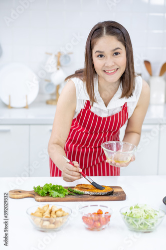 Asian beautiful woman with red apron smear oil and garnish on slicing fish on plate in kitchen with different types of bowl of ingredients on table. Concept of happiness of cooking in their house.