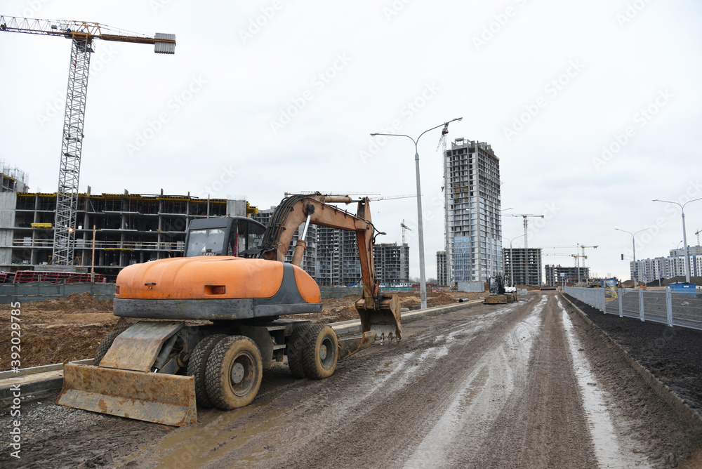 Bucket wheel excavator on road work at construction site. Backhoe digs digs the ground to lay pipes and a new road. Tower cranes are building tall residential buildings.