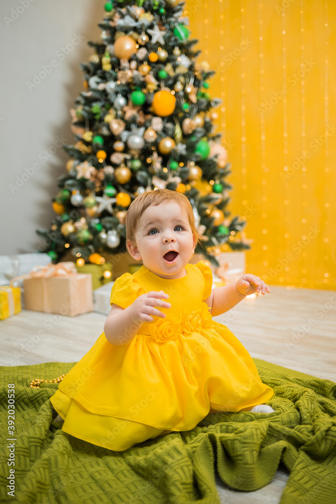 a surprised little girl in a yellow holiday dress sits on the background of a Christmas tree. Vertical orientation