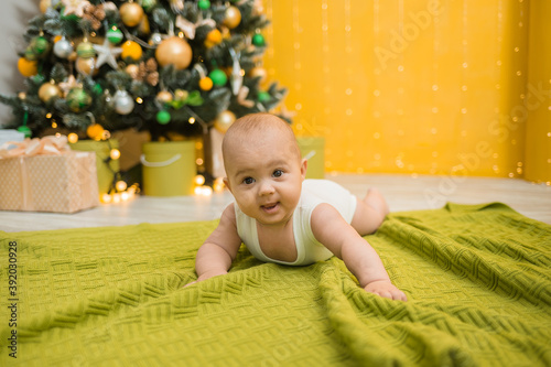 a cheerful little boy in a white bodysuit lies on a green knitted blanket against the background of a Christmas tree