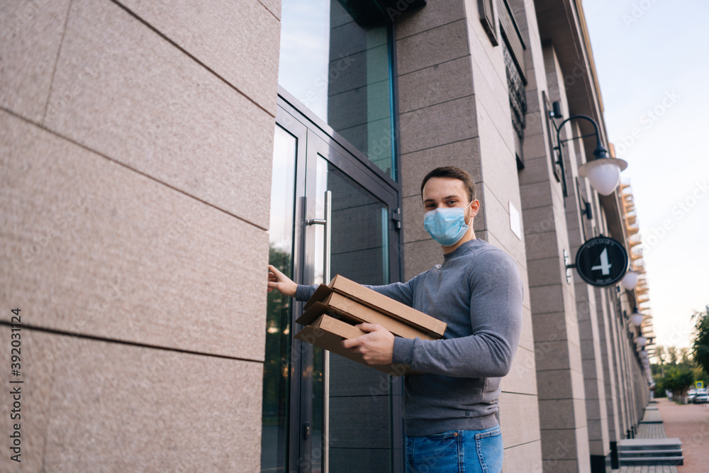 Delivery man wearing medical mask ringing door for delivery carton boxes with hot pizza, looking at camera. Deliveryman in protective mask holding box with food. Concept of delivery during quarantine.