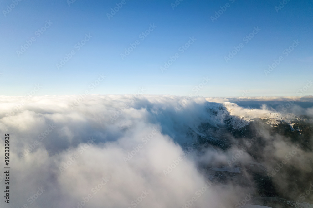 Aerial view from above of white puffy clouds covering snowy mountain tops in bright sunny day.