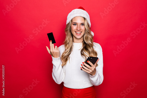 Portrait of a smiling woman in Santa hat holding mobile phone  showing credit card standing isolated over white background