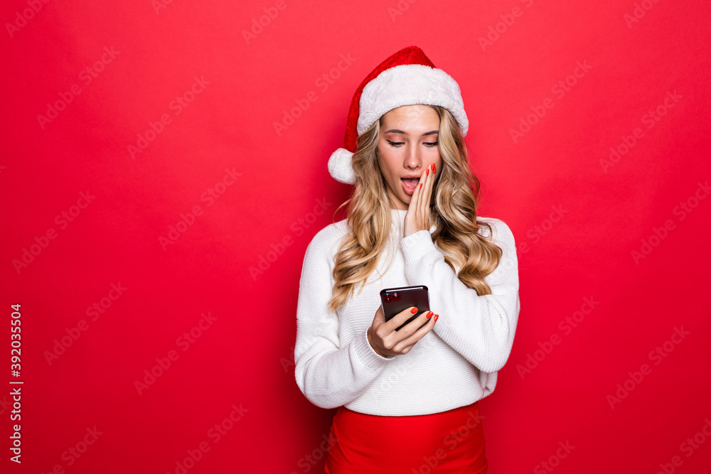 Surprised young woman in santa hat using mobile phone over red background Surprised young woman in santa hat using mobile phone over red background