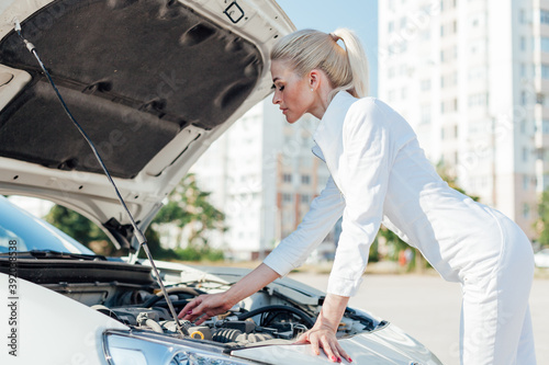 Beautiful female blonde mechanic looks engine under the hood of the car