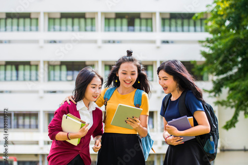 Group of Beautiful Asian students talking together and checking on line content on Tablet with the university building in the background.
