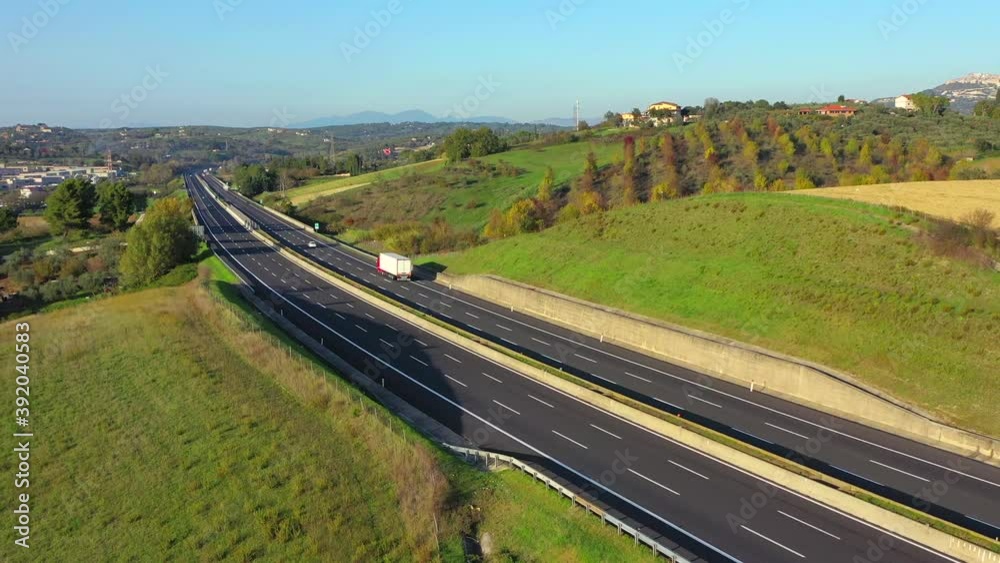 Autostrada a 2 corsie in mezzo alla campagna. Tir, camion, autotreni e auto si sorpassano. Ripresa aerea con drone