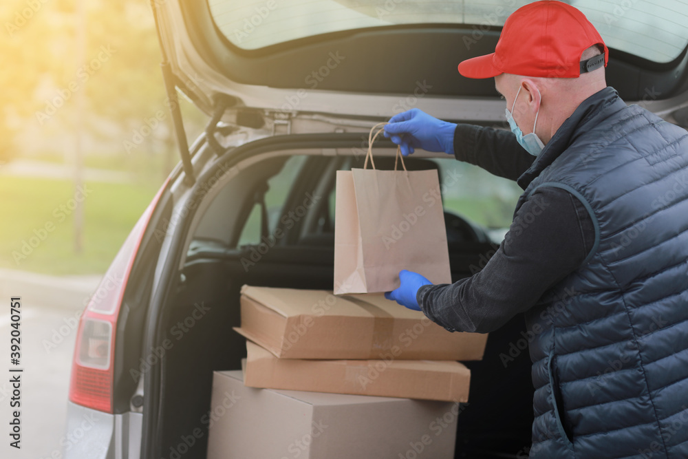 Delivery man in red cap, face medical mask and gloves hold paper bag near the car outdoor. Service coronavirus. Online shopping. mock up. Delivery man in red cap, face medical mask and gloves hold paper bag near the car outdoor. Service coronavirus. Online shopping. mock up.