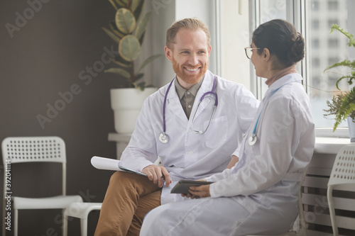 Smiling bearded doctor in white coat talking to young nurse while they sitting on chairs at the corridor