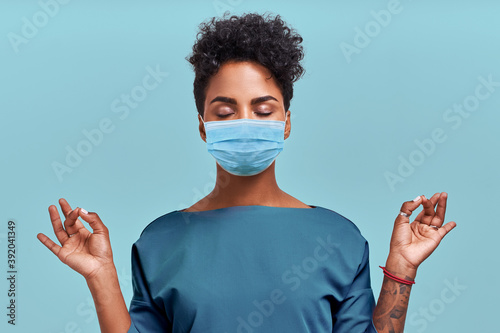 Obraz na plátně African American doctor meditation wear respiratory mask, no stress free relief at work concept, mindful peaceful young female paramedic practicing breathing yoga exercises on blue wall