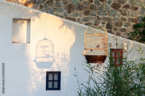 Beautiful atmospheric Mediterranean landscape. Golden cage with goldfinch casts shadow on wall, setting summer sun. Courtyard of house in resort area. Tourism, recreation, relaxation concept