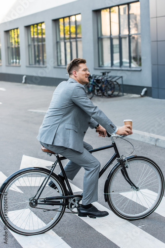 Fototapeta Naklejka Na Ścianę i Meble -  Smiling businessman with coffee to go riding bicycle on crosswalk outdoors