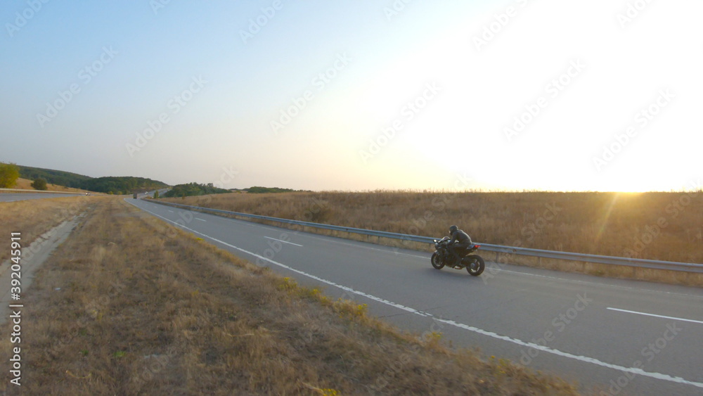 Fototapeta premium Aerial shot of biker is riding on modern sport motorbike at autumn highway. Motorcyclist racing his motorcycle on country road at sunset. Man drive bike during trip. Concept of freedom and adventure