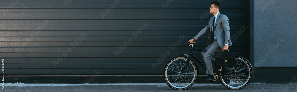 Young businessman holding briefcase while riding bicycle outdoors, banner - obrazy, fototapety, plakaty 