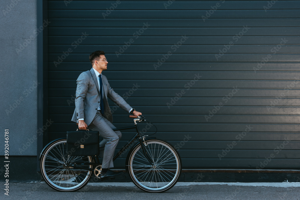 Young businessman with briefcase cycling near facade of building Young businessman with briefcase cycling near facade of building